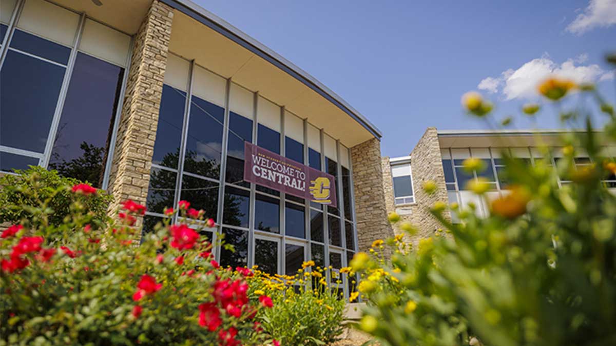Red and white flowers bloom in the foreground of a low angle wide shot of the Bovee University Center.