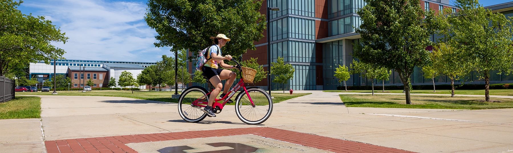 A student wearing a baseball cap and backpack bikes across campus on a sunny day.