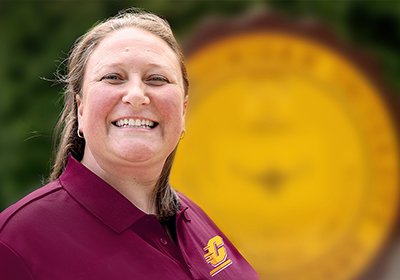 A headshot of Tracy Karr, Assistant Director of Admissions smiling while wearing a maroon shirt and standing in front of the official CMU seal in the Warriner Mall.