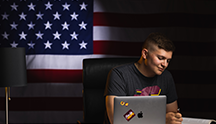 A military student sits at a desk in front of a laptop in a dark room as a well-lit US flag hands from the wall behind them.