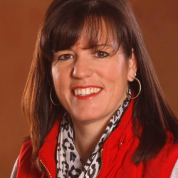 Headshot of Kristina Rouech.  She is standing in front of a copper colored background.  She has long brown hair and gold earrings.  She is wearing a patterned white and brown scarf and a red vest.