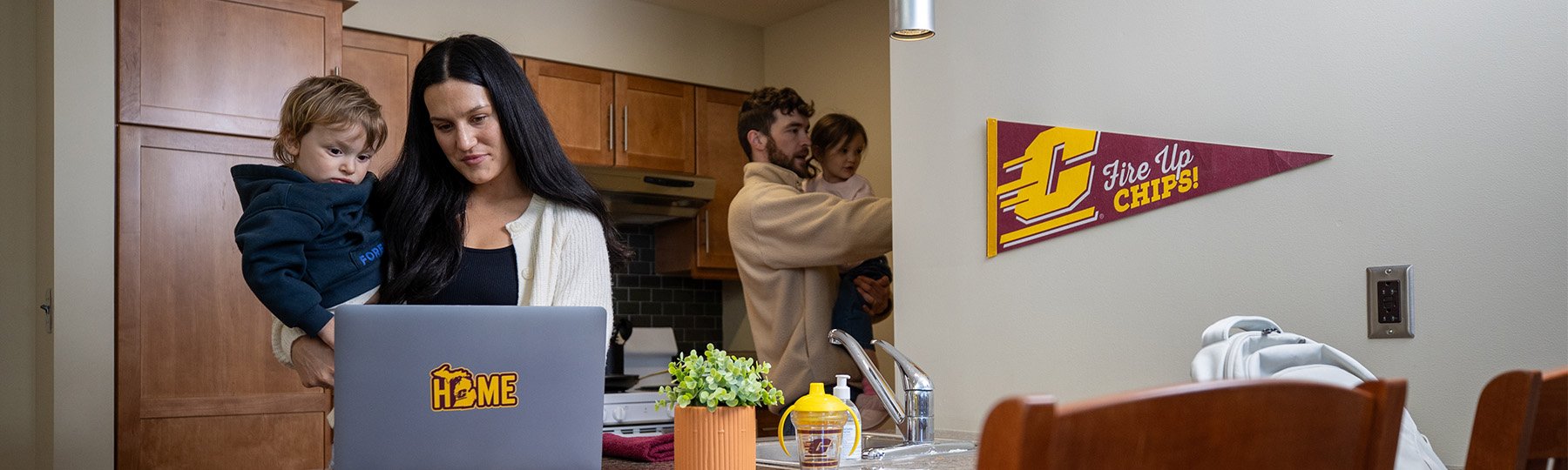 A student works at a laptop in the kitchen with her family in the background.