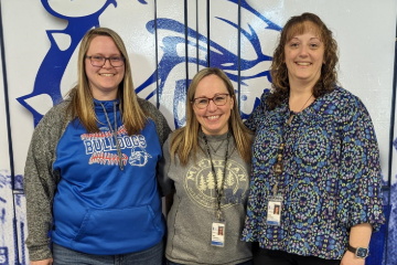 In the background is the Ionia Public School Bulldog Mascot in blue.  In front are three female teachers smiling and posed for the camera.