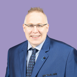 Headshot of Jeremy Hyler on a purple background.  He has very short red hair and wears glasses.  He is smiling and wearing a navy suit with a blue tie.