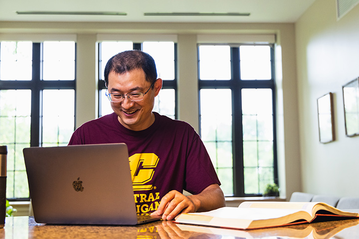A student wearing a CMU T-shirt sits at a table with a laptop and a text book. The student is smiling at his laptop. There are windows behind him.