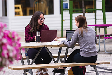 Two students sitting at a picnic table with their laptop computers. One student is facing the camera and smiling. There are pink flowers in the foreground.