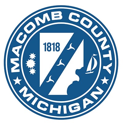 A blue and white circular logo with the words Macomb County Michigan in white in the center is a shape of the county in white and the number 1818 in blue. On each side of the county are three gears and a sailboat.