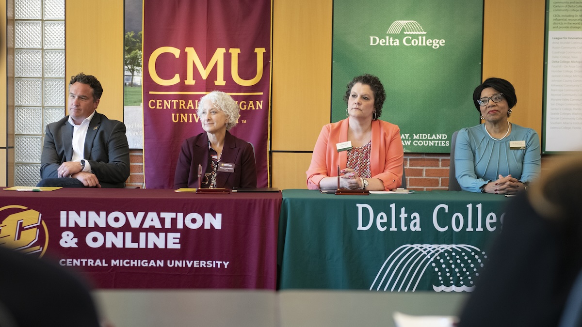 social-partnership-cmu-delta-college-20260623.01 A table with a Central Michigan University table cover sits next to a table with a Delta College table cover. One man and three women wearing business attire sit behind the tables.