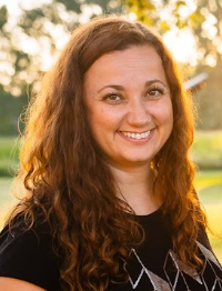 Headshot of Margo Wilson outside on a bright sunny day.  There is a tree behind her and bright skies.  She has long curly brown hair and a big smile.  She is wearing a black tshirt with a geometric pattern on the front