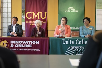 A table with a Central Michigan University table cover sits next to a table with a Delta College table cover. One man and three women wearing business attire sit behind the tables.