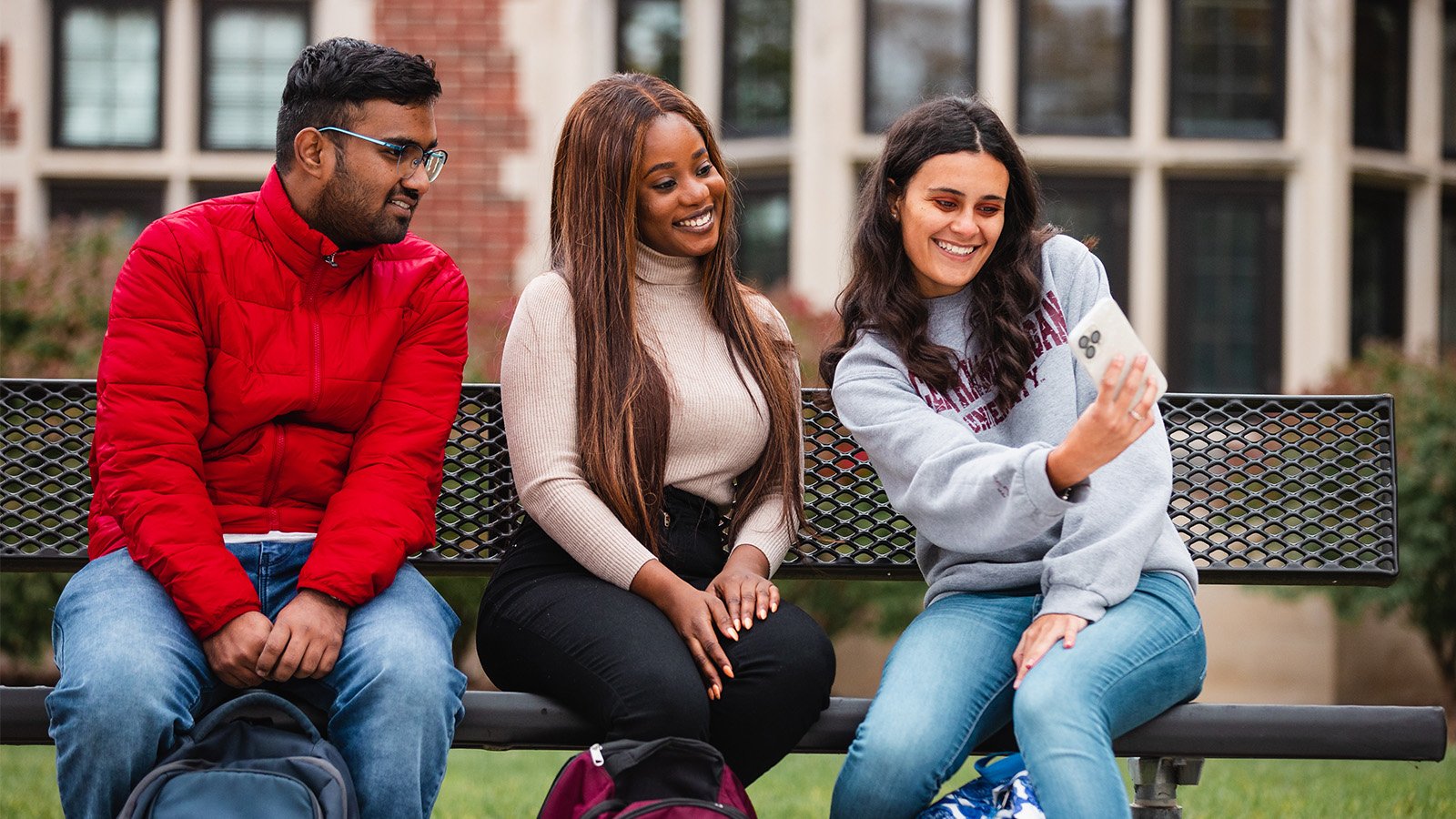 2021-187-148 International students stock_1600x900 Three students sit on a campus bench together, smiling and posing for a selfie-style photo