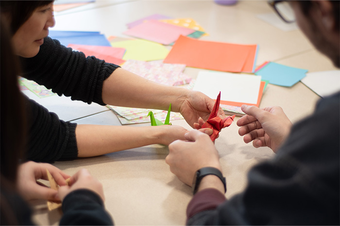 Two individuals work together to fold an origami paper crane. A close up of their hands.