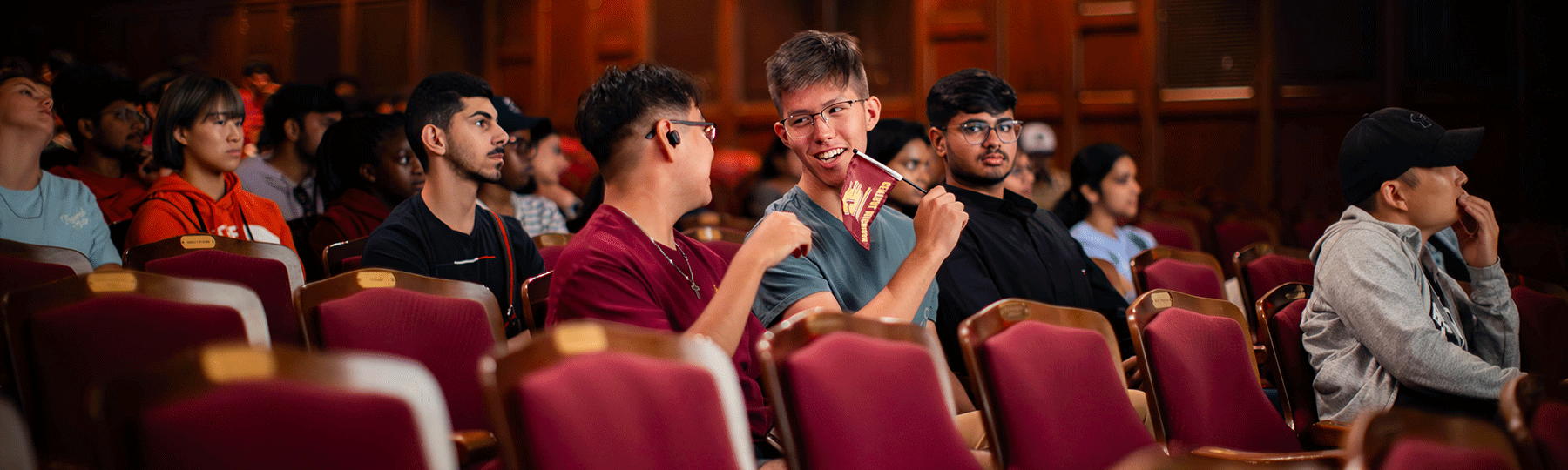 Students sitting in an auditorium while listening to speakers present for the international student orientation. One male student is holding a small maroon CMU flag with a gold action C on it.