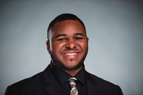 Professional headshot of a smiling Donovan Pitts-Reed wearing a black jackets, black shirt, and a plaid tie against a light grey backgound.
