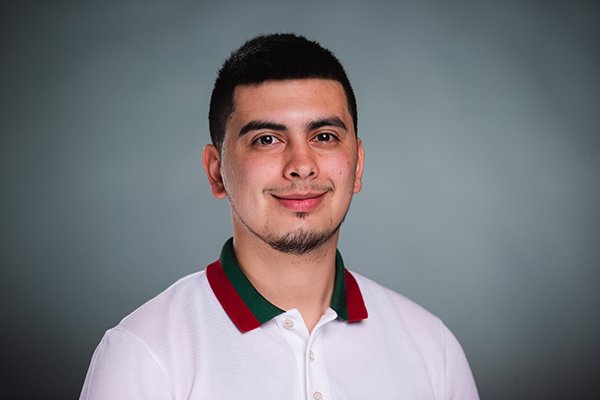 Professional headshot of a smiling Marcelino Olivarez wearing a white shirt with red and black collar against a light grey backgound.
