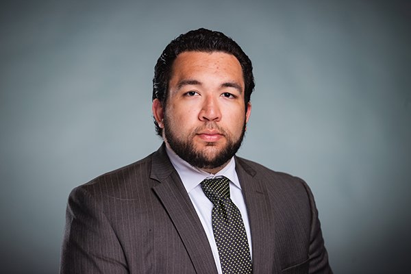 Professional headshot of Max Logan wearing a grey pinstriped jacket, white shirt, and black tie with small yellow polka dots against a light grey backgound.