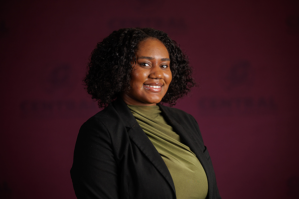 Professional headshot of Arterah Griggs wearing a green shirt with black blazer on a maroon background.