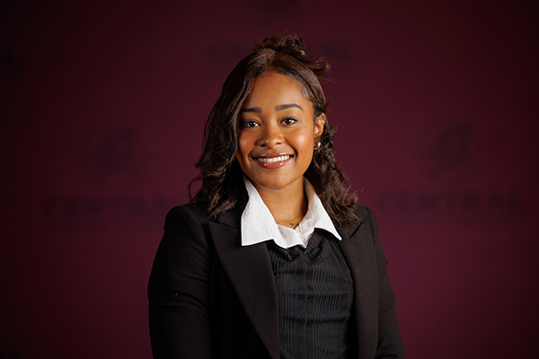 Professional headshot of Mireya Benjamin wearing a white shirt under a black sweater and blazer on a maroon background.