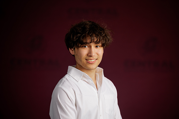 Professional headshot of Nicholas Spicer wearing white button-up shirt on a maroon background.