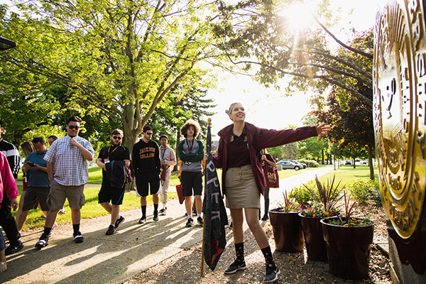 A female orientation leader in maroon and gold leads a group of students on a campus tour near the Central Michigan University seal in Warriner Mall