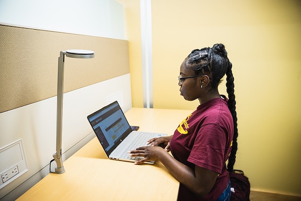 A female student in a maroon t-shirt sits at her laptop in a stark, yellow room