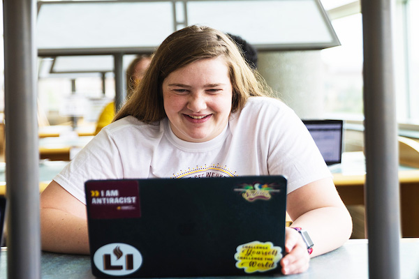 A female student smiles as she faces the camera and works on laptop covered with stickers.