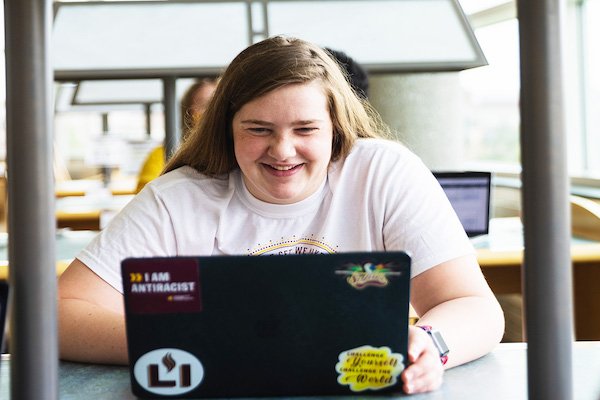 A female student smiles as she faces the camera and works on laptop covered with stickers.