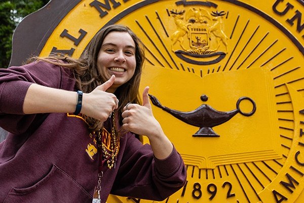 A student with a maroon CMU sweatshirt on with her thumbs up in front of the CMU seal