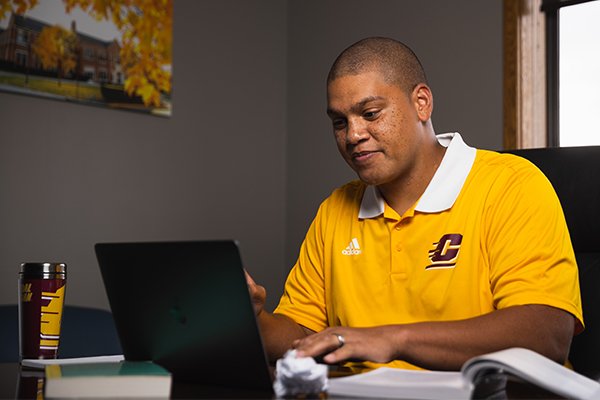 a student with a gold CMU polo shirt on sitting at a desk using a laptop