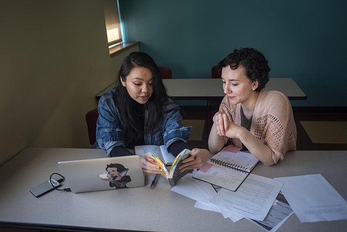 Two students sitting at a table with laptop looking at a book together