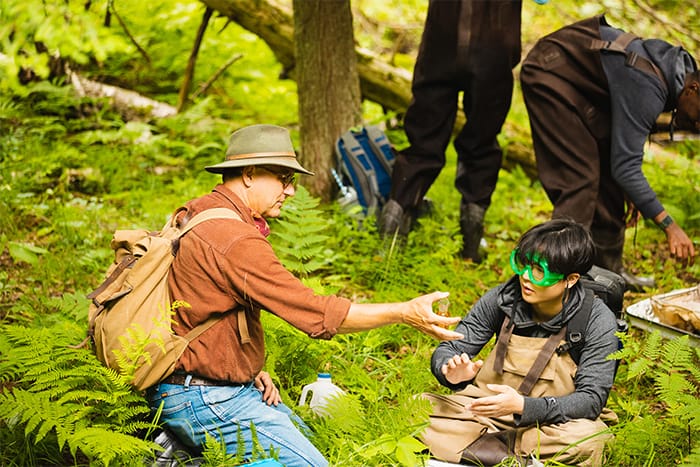 A professor wearing an orange shirt, backpack and hat shows a student wearing safety google and waiters a sample.
