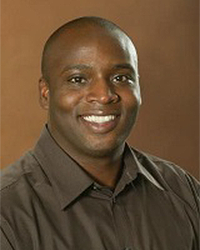Adrian Lamar is an Alumni Board member who holds Emeritus status and he is standing in front of a light brown background wearing a dark brown shirt.