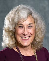 Pam Murray is an Advancement Board member and she is wearing a maroon top and a maroon and gold necklace and she has shoulder-length curly hair.