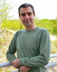 A man wearing a long-sleeved turquoise shirt man leans on a railing with green foliage behind him.