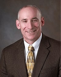 Bret Hyble is an Alumni Board member and he is wearing a brown suit with a yellow tie with a pattern and a white button down shirt.