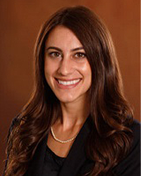 Brittany Mouzourakis is an Alumni Board member who has Emeritus status and she has long dark brown hair and is wearing a black top and pearls.