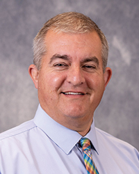 Charles Selinger is an Alumni Board member who holds Emeritus status and he is wearing a maroon white shirt with a colorful tie.
