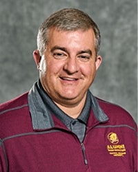 Charles Selinger is an Alumni Board member who holds Emeritus status and he is wearing a maroon alumni shirt with the Action "C" and has short hair.