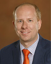 Chris Gautz is an Alumni Board member and he is wearing a navy suit with an orange tie and he has short hair.