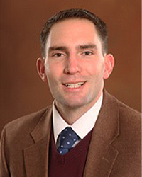 Derek Berger is an Alumni Board member who holds Emeritus Status and is wearing a brown suit, a blue tie with white polka dots, and a dark brown vest and he has short brown hair.