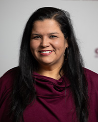 Headshot of Alumni Board of Directors Emeritus Member Rebeca Barrios-Hurst who has long black hair and is wearing a maroon shirt in front of an Alumni Relations background.