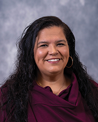 Headshot of Alumni Board of Directors Emeritus Member Rebeca Barrios-Hurst who has long black hair and is wearing a maroon shirt in front of a portrait background.