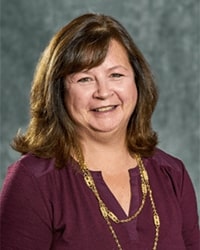 Alumni Board of Directors member Jacalyn Goforth wearing a dark maroon shirt and golden necklace and she has shoulder-length light brown hair in front of a gray background.