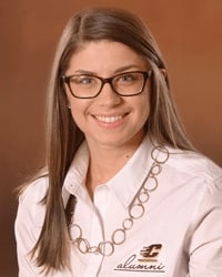 Jaime Leyrer is an Alumni Board member who holds Emeritus status and she is wearing a white alumni shirt with the Action "C" and has long light brown hair and is wearing glasses.