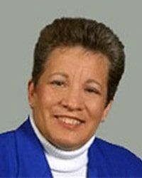 Janet Burns is an Alumni Board member who holds Emeritus status and she is wearing a white turtleneck top and a bright blue suit jacket and she has short brown hair.