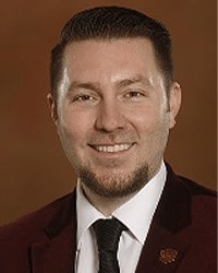 James J. (JJ) Lewis is an Alumni Board member and he has short brown hair and a beard and is wearing a maroon suit jacket and black tie.
