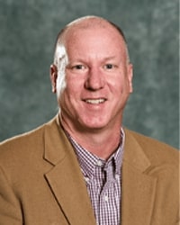 Alumni Board member Jonathan Eadie standing in front of a gray background and he is wearing a tan suit jacket and a maroon plaid shirt.