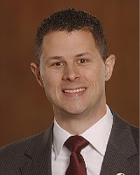 Joshua Richards is an Alumni Board member who holds Emeritus status and he has short curly brown hair and is wearing a gray suit with a maroon tie.