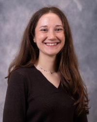 Headshot of Alumni Association Board of Directors member Jennifer Peacock wearing a light black shirt and she has long curly brown hair and is smiling with teeth.