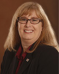 Kandra Robbins is an Alumni Board member and the current President and she is wearing a black blazer with a maroon top and she has shoulder-length red hair and glasses.